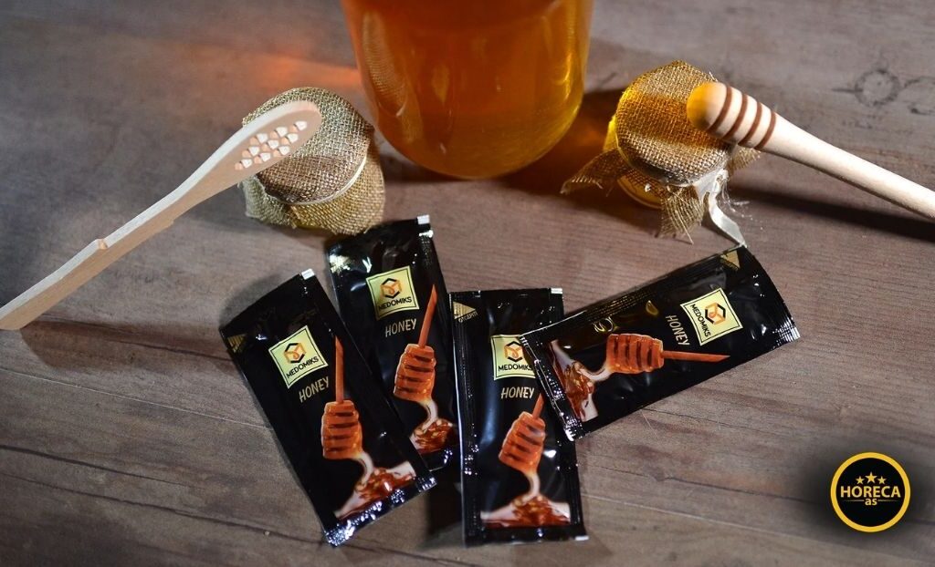Pojedinačno pakovane kesice meda pored tegle meda i drvene kašike na stolu