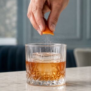 Istiskivanje ulja iz kore pomorandže iznad Old Fashioned koktela sa velikom kockom leda.
